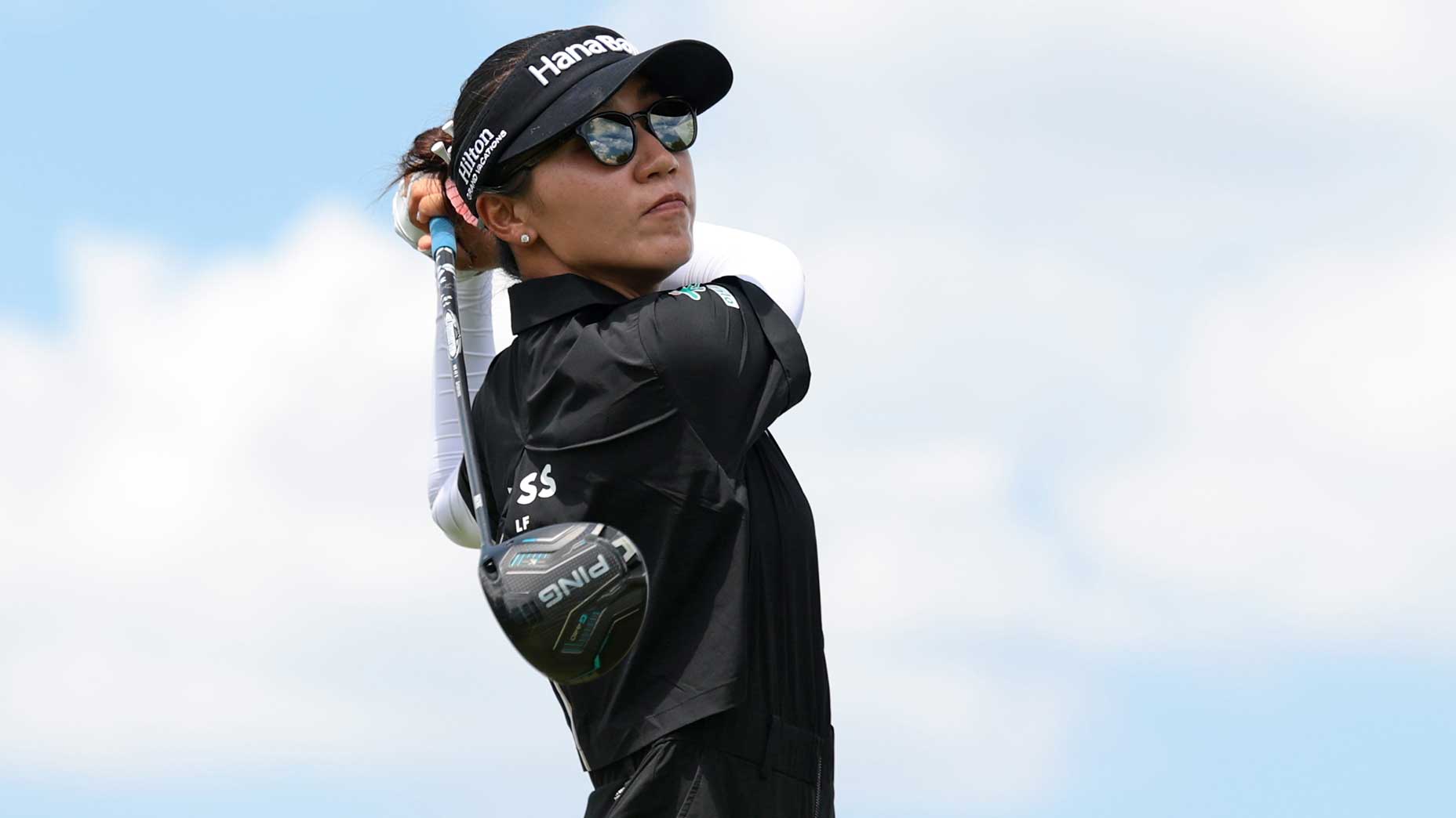 A female golfer, reminiscent of Nelly Korda, wearing sunglasses, a black cap, and black outfit swings a golf club during a game under a partly cloudy sky.