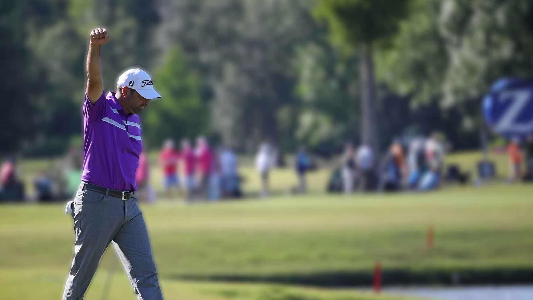 Marc Turnesa wearing a purple shirt, gray pants, and a white cap raises his fist in celebration at the U.S. Open, with blurred spectators and trees captured by a Sony camcorder in the background.