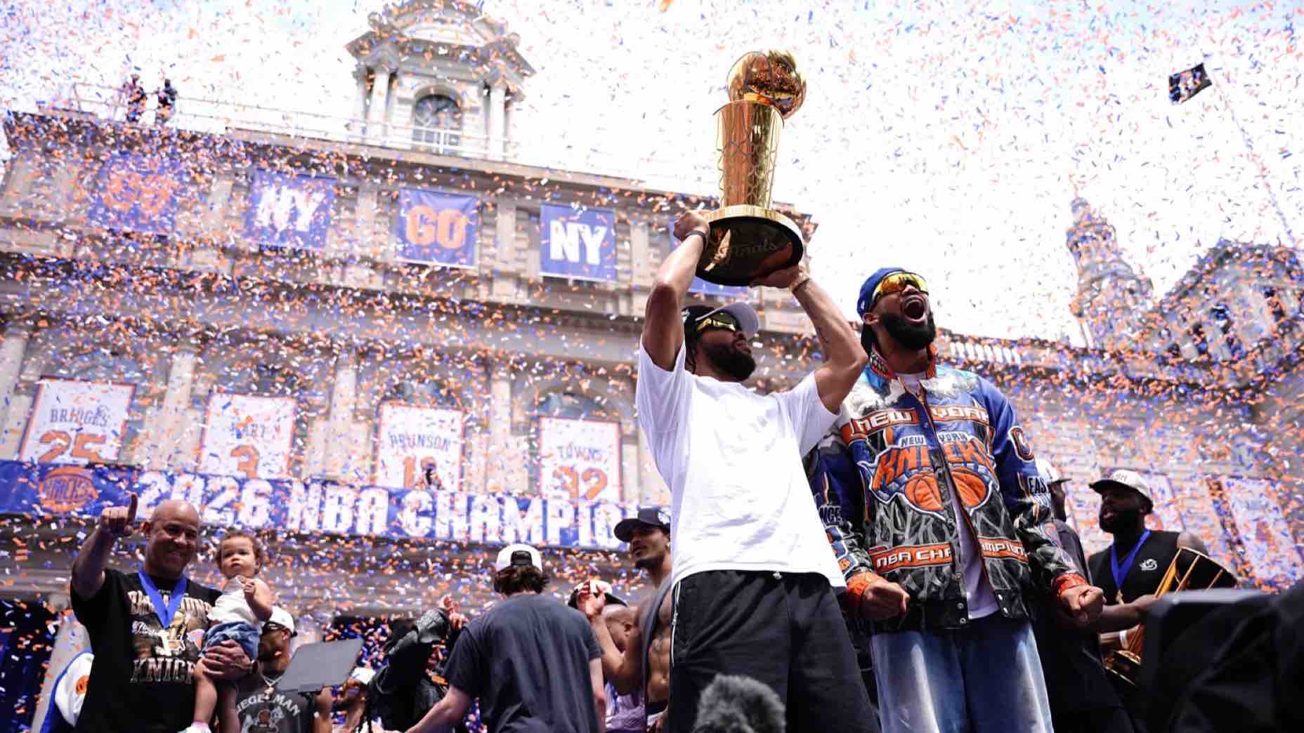 A basketball player holds up the NBA championship trophy amid falling confetti, surrounded by celebrating teammates and fans. Banners read 2026 NBA CHAMPIONS in New York Knicks colors, marking a highlight in New York sports during the greatest sports week.