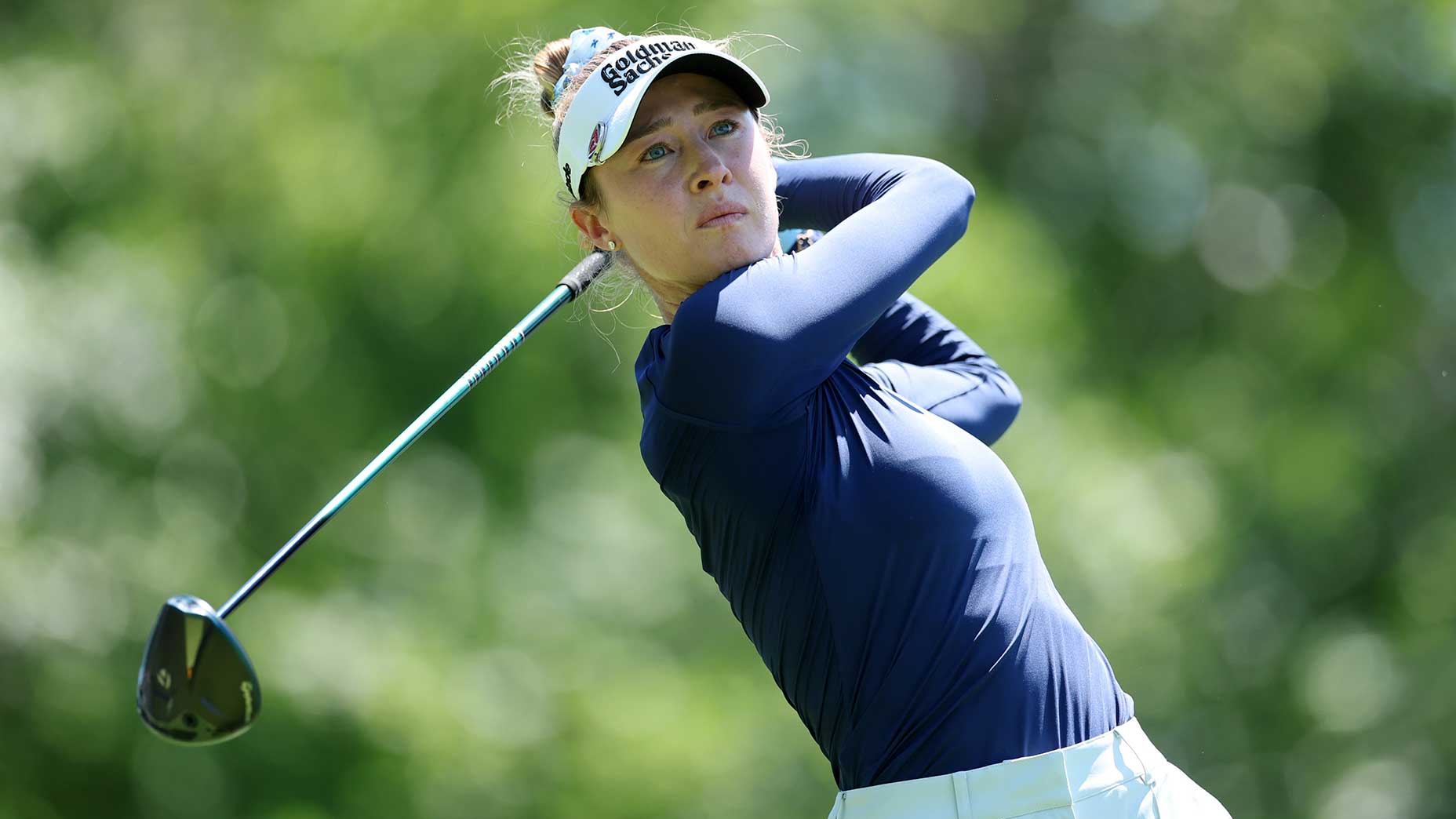 A female golfer in a navy blue long-sleeve shirt and white visor follows through on her swing, channeling Nelly Korda's historic streak at the KPMG Women's PGA, with greenery blurred in the background.