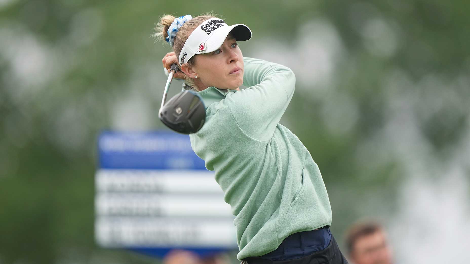 A female golfer, reminiscent of Nelly Korda, wearing a light green jacket and visor swings a golf club during a game at the KPMG Women's PGA, with a blurred scoreboard and trees in the background.