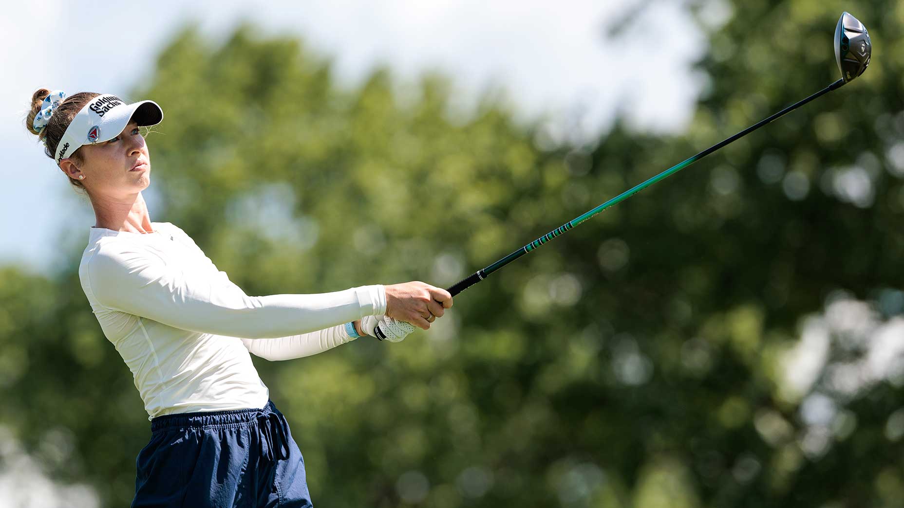 A female golfer wearing a visor, long-sleeve shirt, and dark pants watches her shot after swinging a golf club, with trees and blue sky in the background.