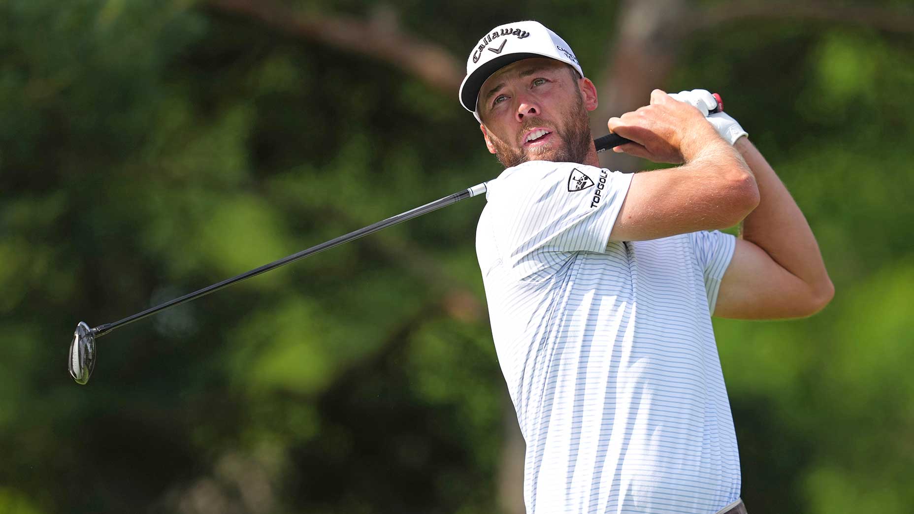 A male golfer wearing a white cap and striped shirt follows through on a golf swing, holding a club over his shoulder with trees blurred in the background during Saturday tee times at the Memorial Tournament.