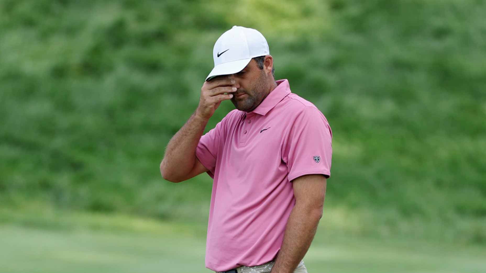 A golfer in a pink shirt and white hat stands on the course with his hand covering his face, appearing frustrated or disappointed, with a blurred green background.