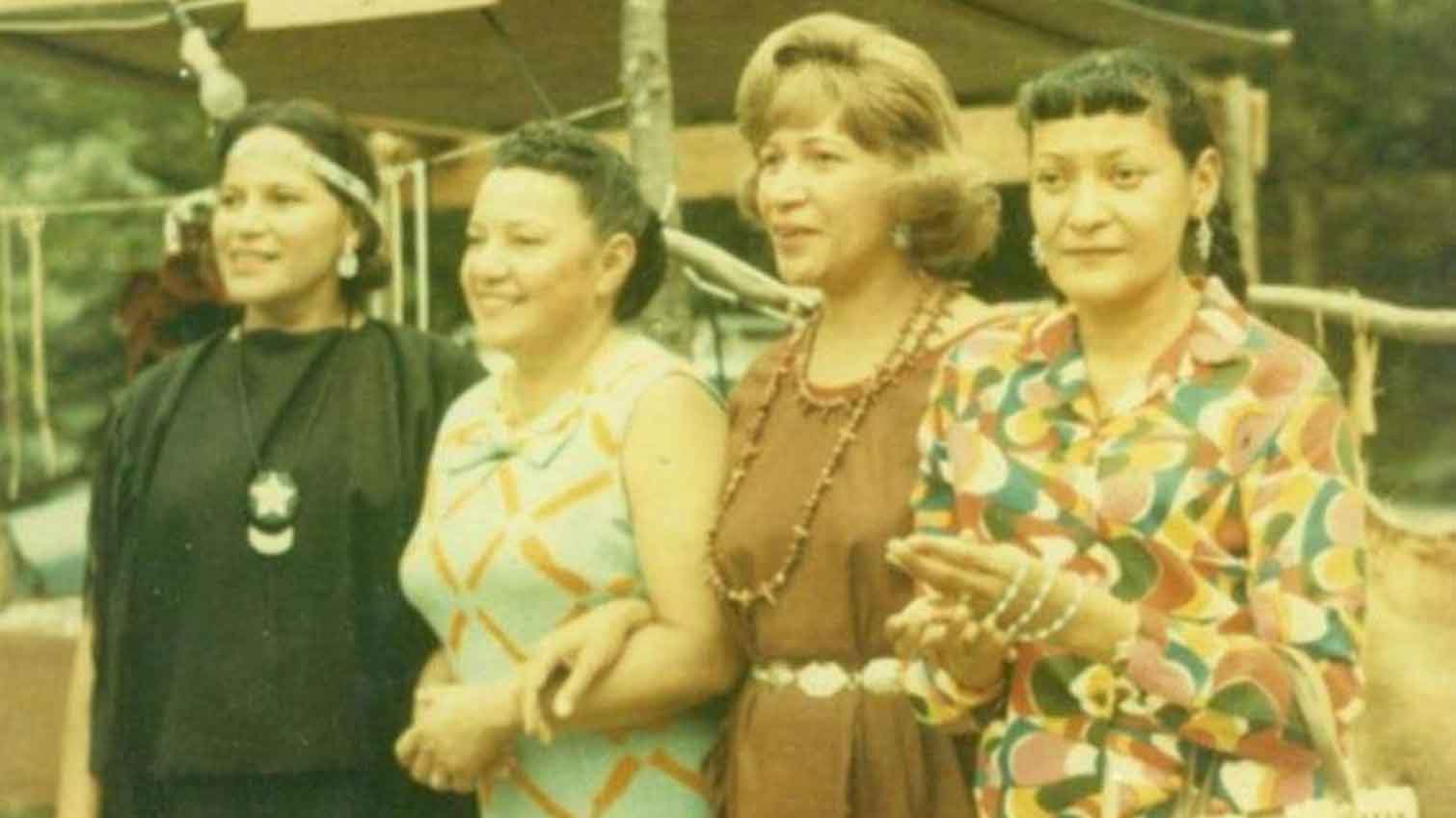 Four women stand closely together outdoors in Shinnecock Hills, smiling and holding hands. Dressed in colorful, patterned clothing and jewelry, they share a family bond. Trees and a wooden structure appear in the vintage photo’s background.