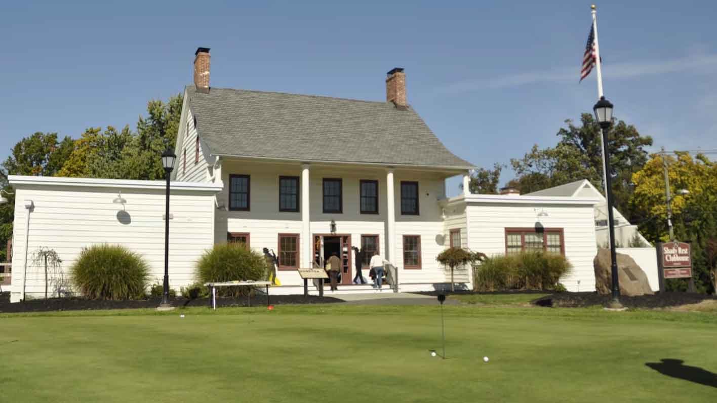 A white colonial-style building with columns, two chimneys, and people entering sits behind a green lawn with a putting area—a nod to the U.S. Open; an American flag flies on a tall pole to the right.