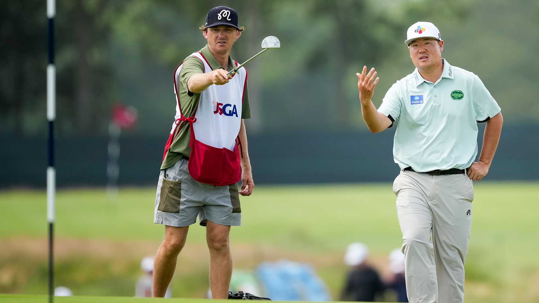 A golfer in a light green shirt talks with his caddie, who points towards the green while holding a golf club. They are standing on a golf course near the hole, with trees and blurred figures in the background.