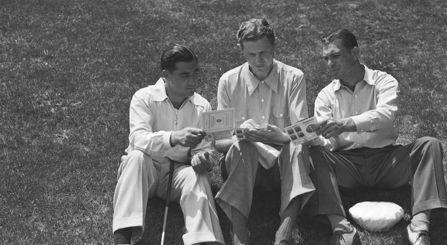 Three men sitting on grass, dressed in long-sleeved shirts and trousers, using a Sony camcorder while discussing papers or booklets together on a sunny day.