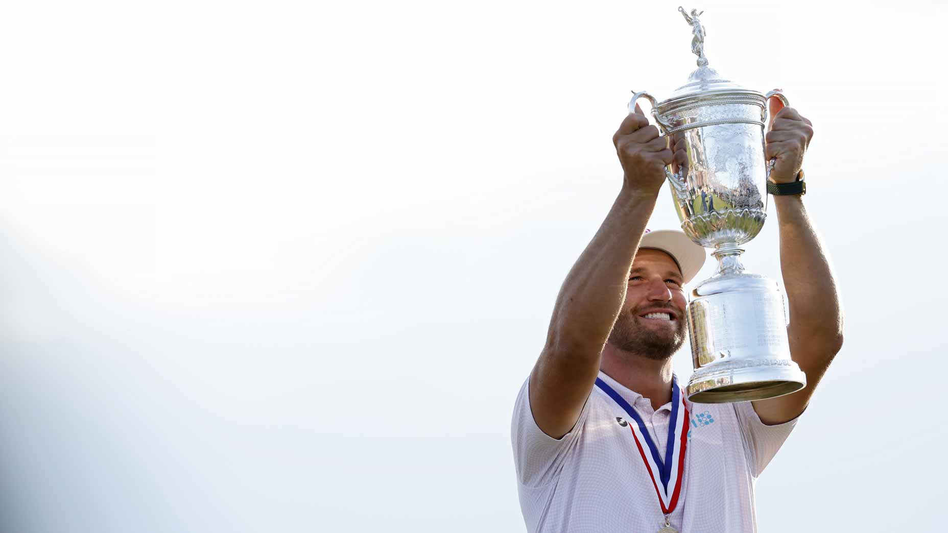 Golfer Wyndham Clark, wearing a white shirt and cap, smiles while lifting a large silver U.S. Open trophy overhead, with a medal around his neck, standing against a bright, white background.