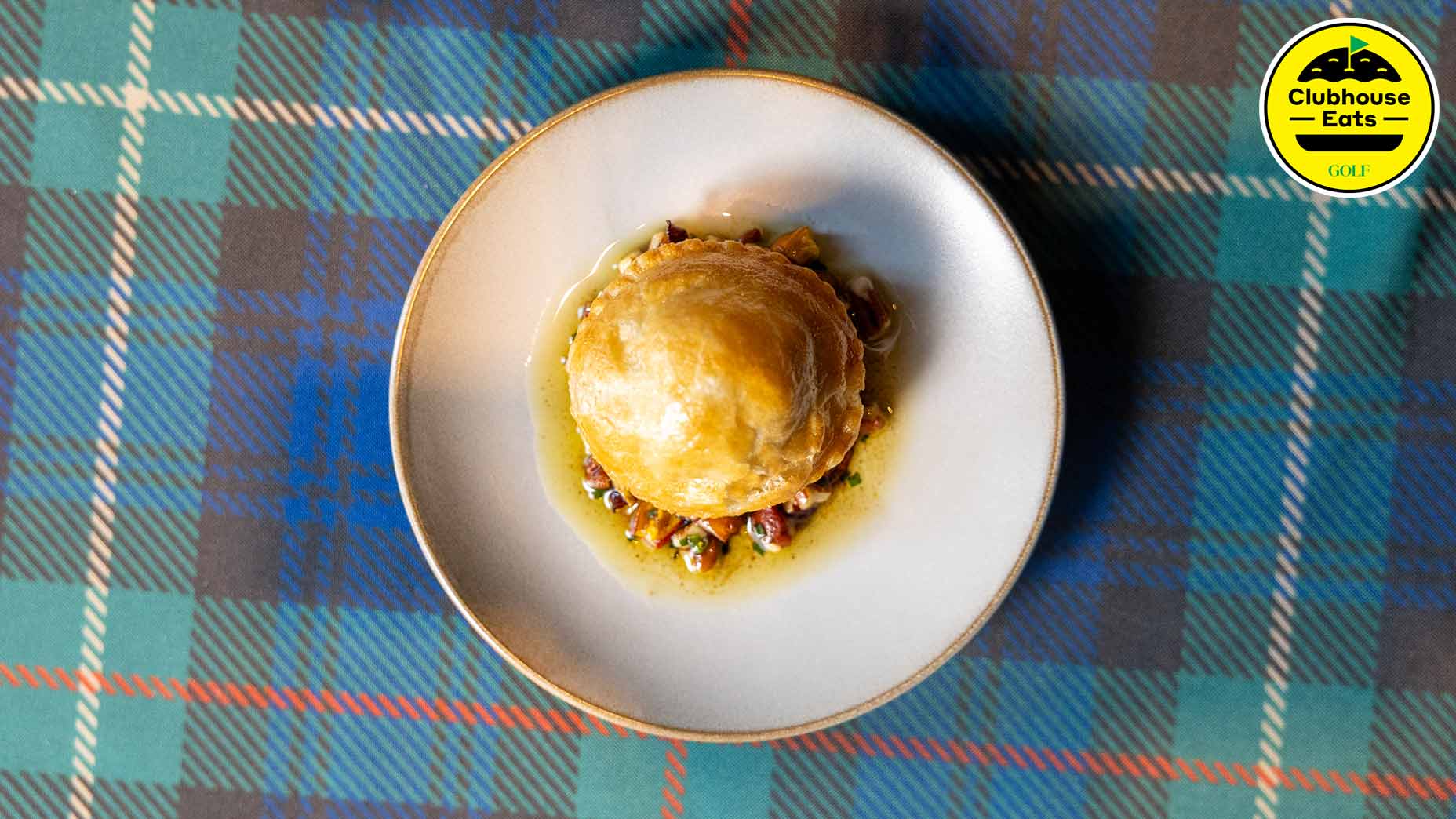 A round puff pastry sits on a plate with sauce and vegetables, reminiscent of the best bread pudding, placed on a blue plaid tablecloth. A “Clubhouse Eats” logo is in the top right corner.
