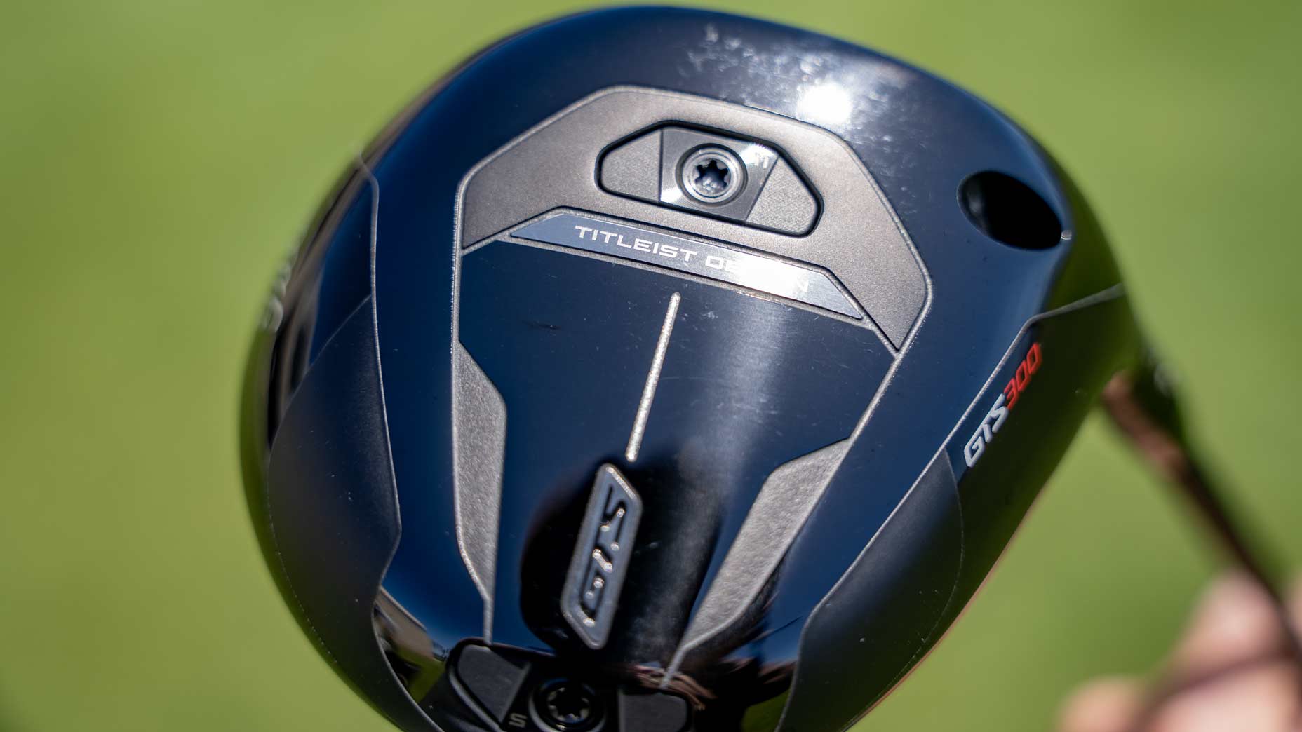 Close-up of a black Titleist GTS300 golf club head with “TS2” and “Titleist” branding, screws, and sleek silver accents, highlighting the Mini Driver design, set against a blurred green background.