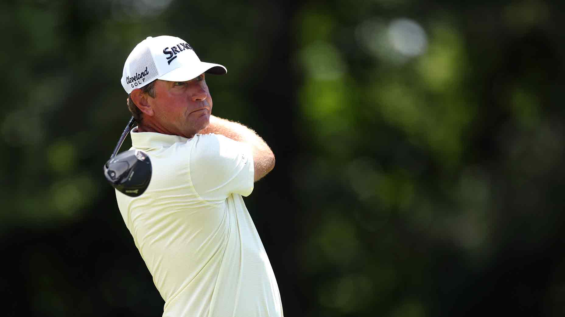 A golfer wearing a white shirt and cap swings a golf club at the John Deere Classic, with green blurred trees in the background.