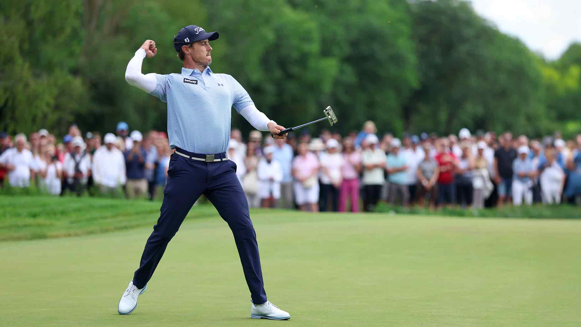 A golfer in a blue and white outfit throws his putter in frustration on the green during a DP World Tour event, with a large crowd of spectators watching in the background.