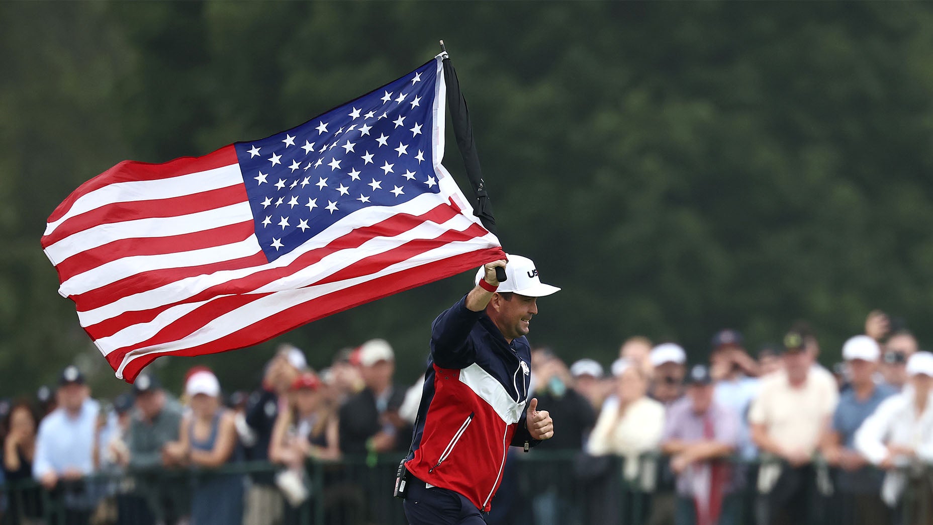 A golfer in a red, white, and blue jacket waves a large American flag while walking on a golf course, with a crowd of spectators blurred in the background.