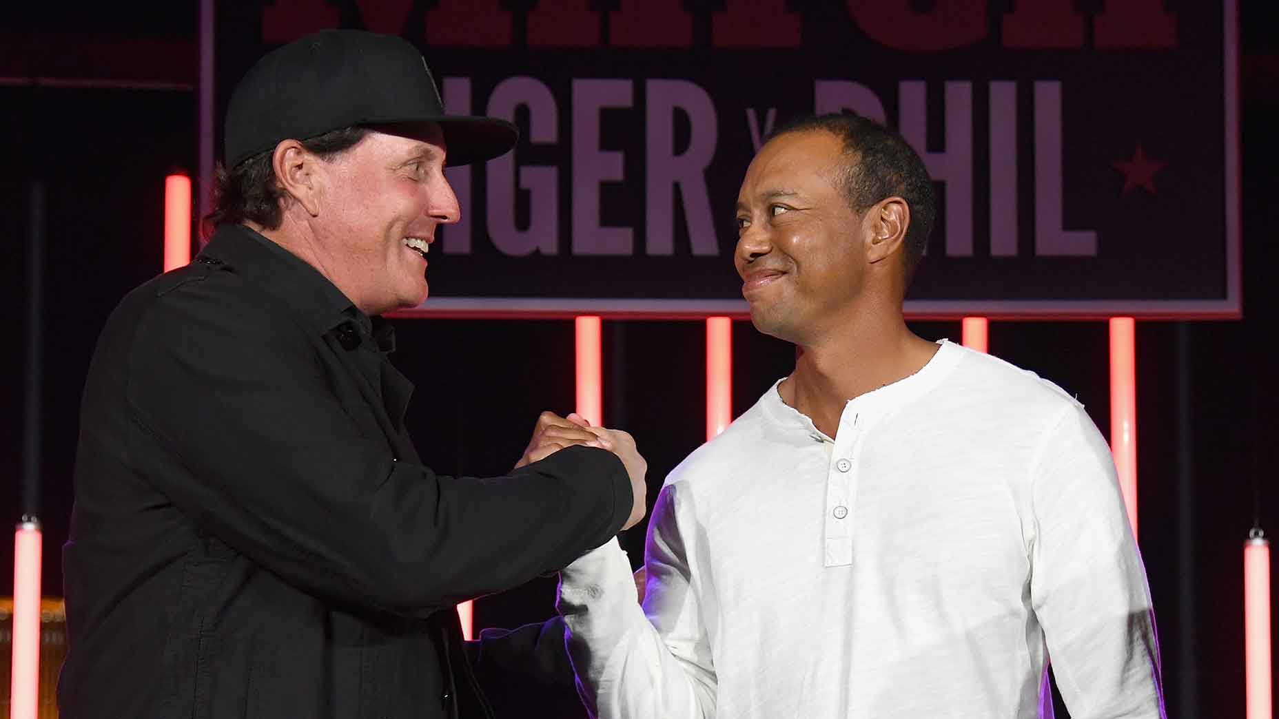 phil mickelson and tiger woods smiling and shaking hands in front of a sign with bold letters. One wears a black jacket and cap, the other wears a white long-sleeve shirt. Neon lights and a dark background create a friendly, casual atmosphere.