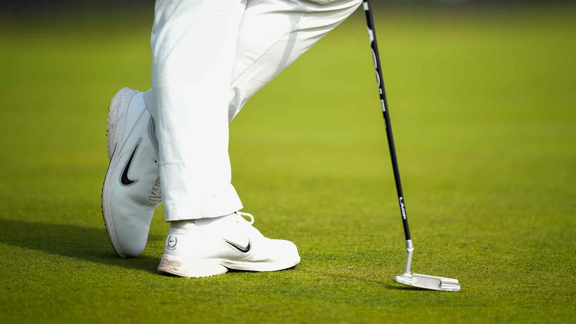 A golfer wearing white pants and white Nike golf shoes, reminiscent of Tom Kim’s signature style, stands on green grass, holding a putter with the head resting on the ground.