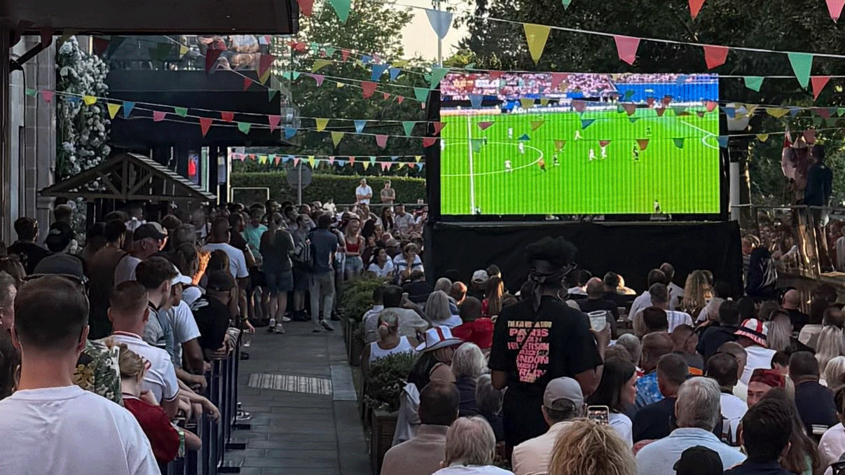 A large crowd watches a soccer match on a big outdoor screen, with colorful bunting overhead and people sitting and standing closely together.