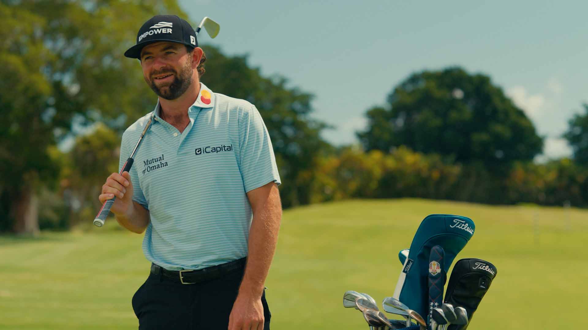 A smiling golfer in a striped polo shirt and cap stands on a golf course holding a club, with a golf bag full of clubs beside him. Trees and green grass are in the background.