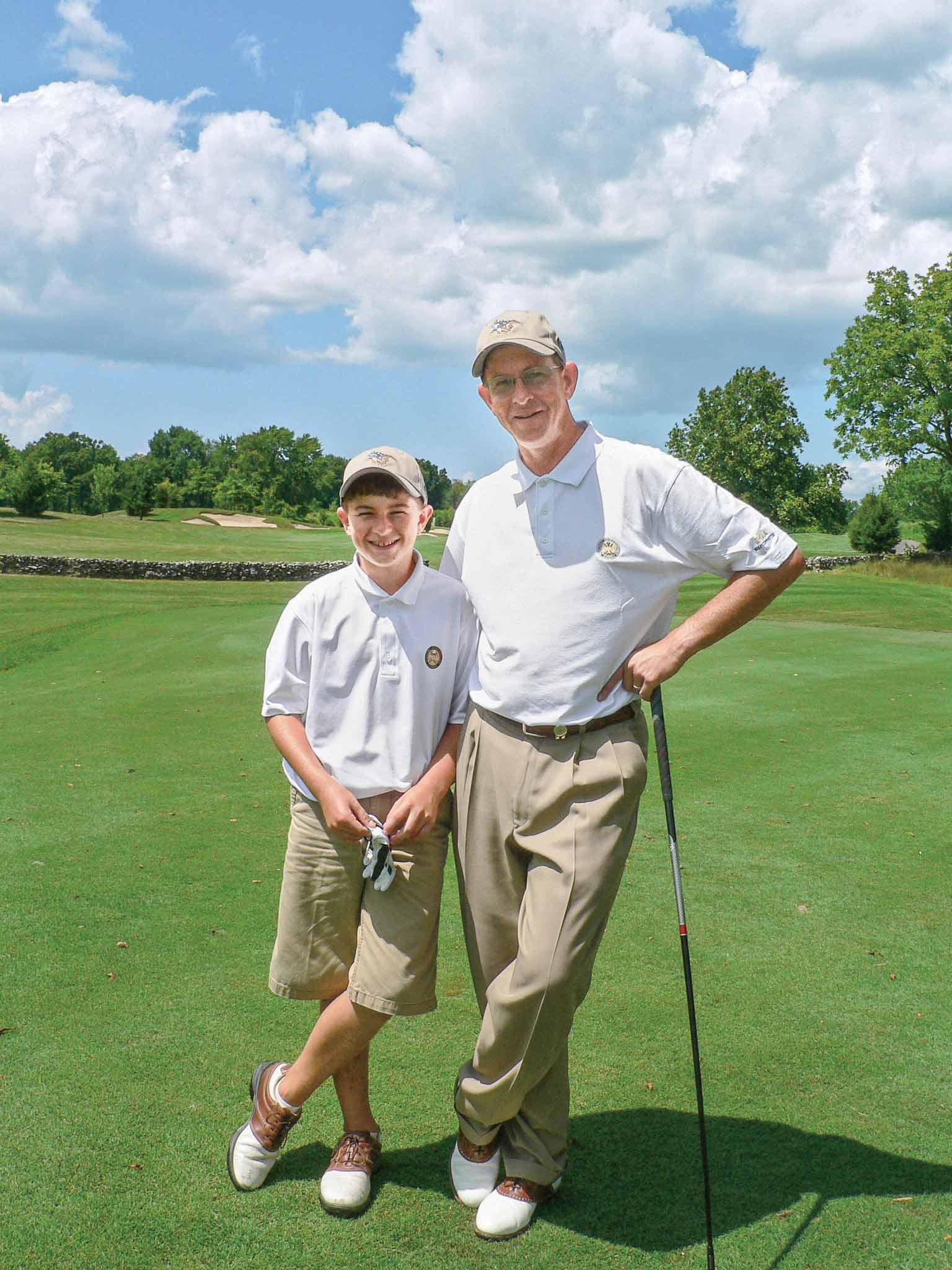 A man and a boy wearing golf attire stand together on a golf course, smiling at the camera. The man holds a golf club and both are dressed in white shirts, khaki pants, and caps. The sky is bright and partly cloudy.