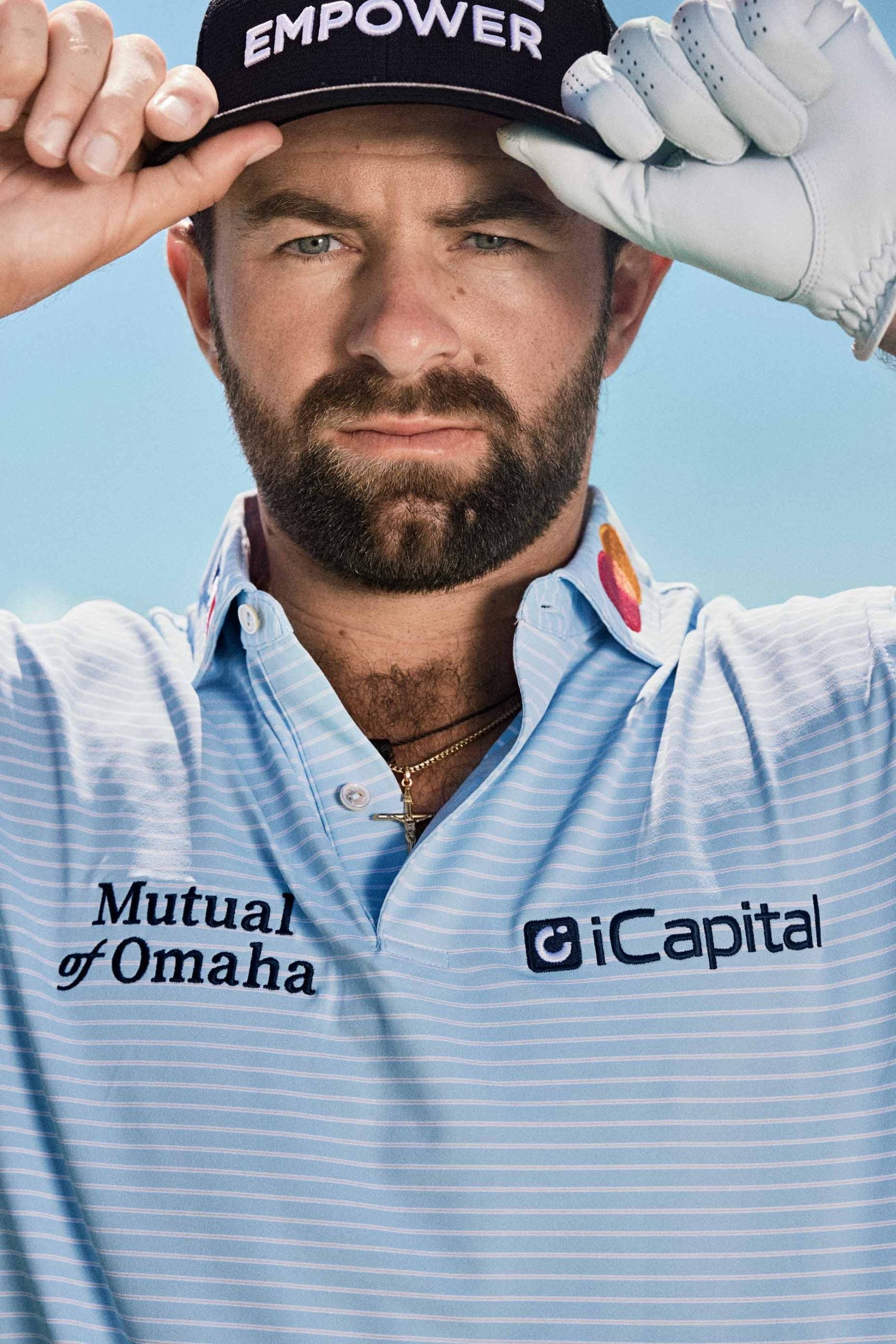 A man wearing a golf glove and a dark cap that says EMPOWER lifts the brim of his hat. He has a beard and is dressed in a light blue striped golf shirt with Mutual of Omaha and iCapital logos.