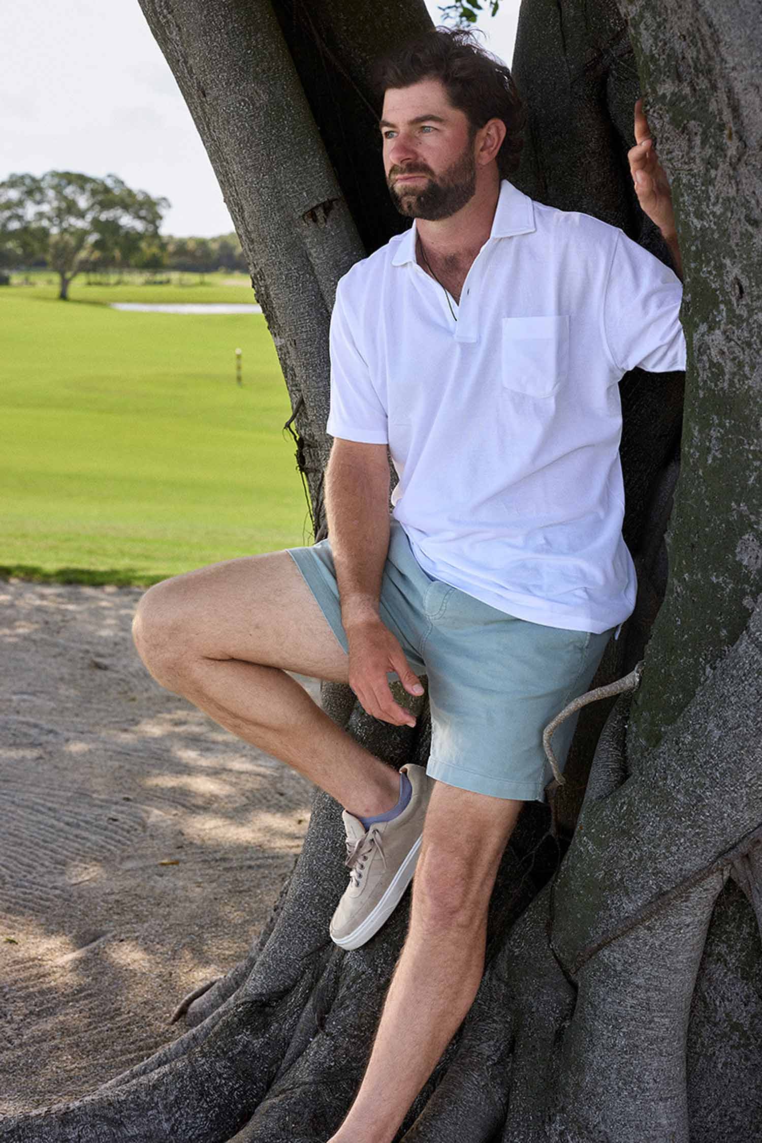 A man with a beard sits on a large tree root, wearing a white polo shirt, light blue shorts, and sneakers. A grassy field and distant trees are visible in the background.