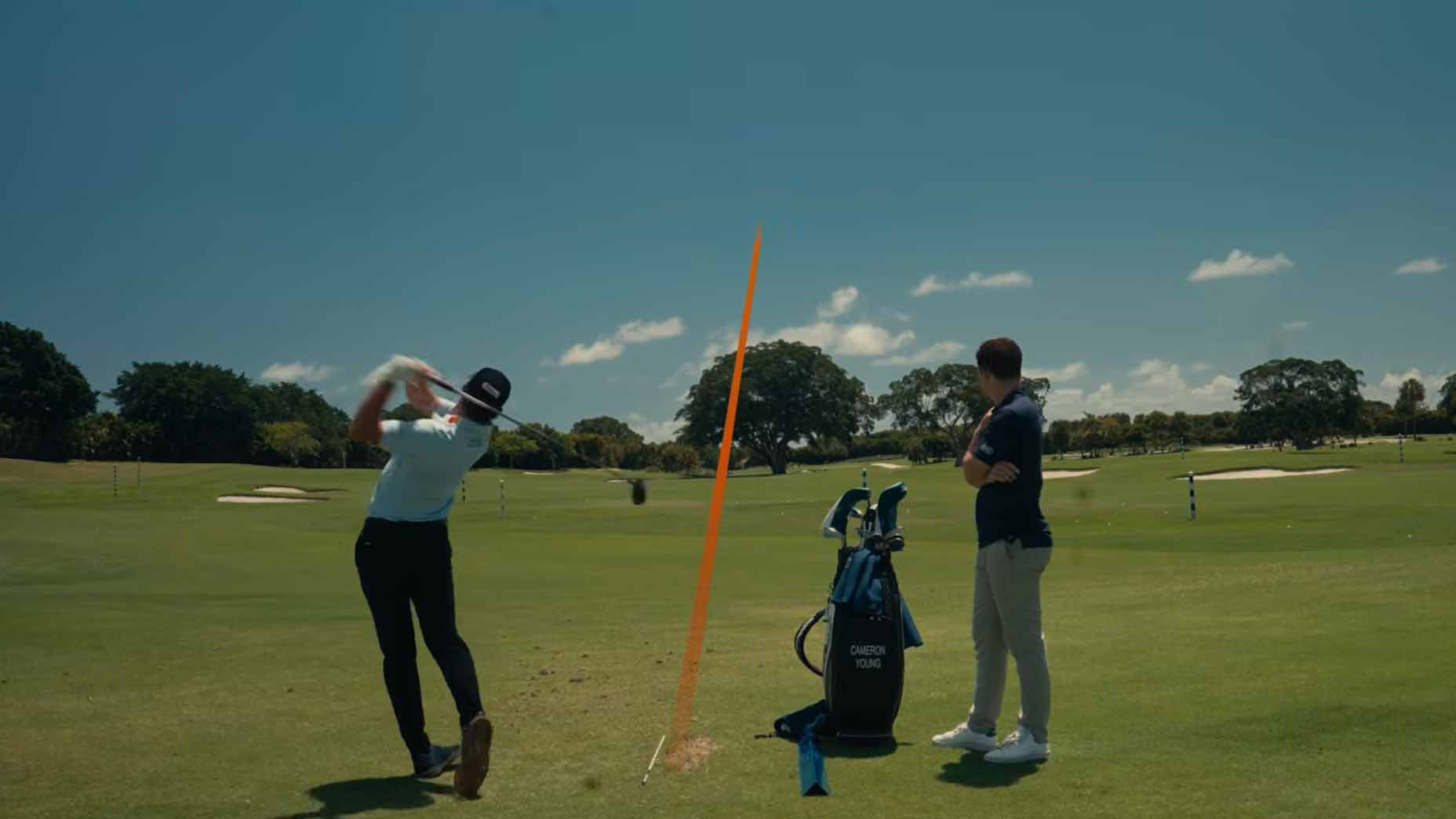 A golfer swings a club as another person watches on a sunny golf course. An orange line shows the balls trajectory. A golf bag sits nearby.