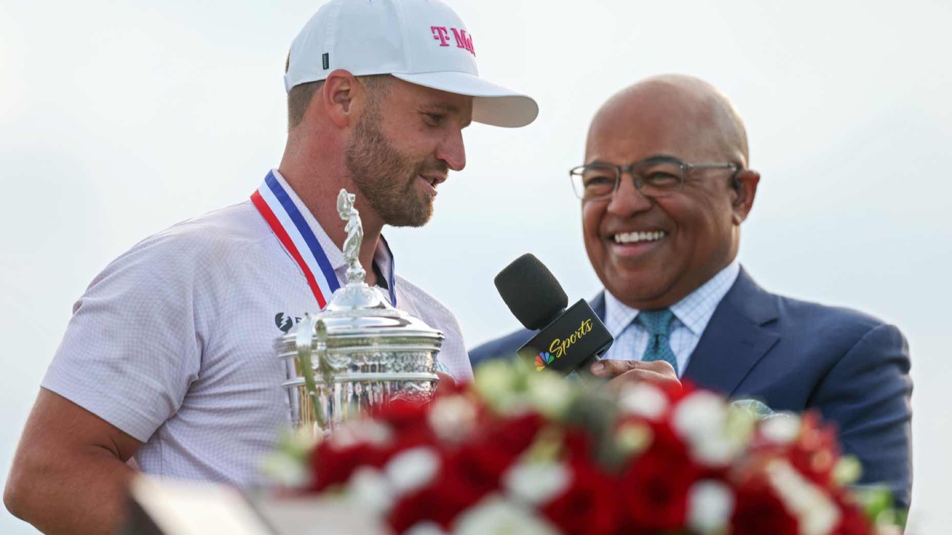 A golfer wearing a medal holds a trophy while being interviewed by a smiling broadcaster in a suit holding a microphone. A bouquet of red and white flowers is visible in the foreground.