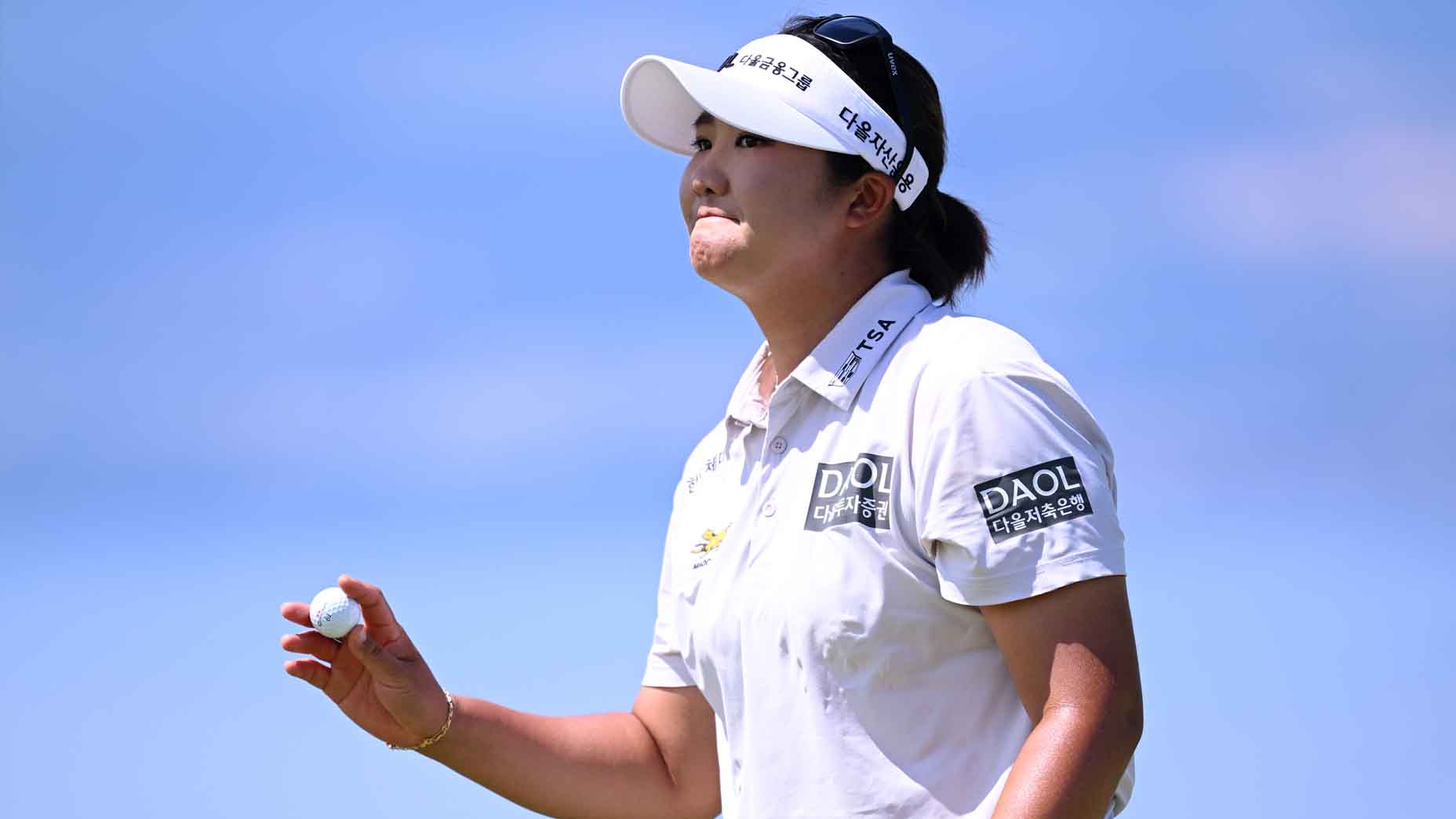 A golfer wearing a white shirt, cap, and sunglasses holds a golf ball in one hand, looking ahead with a focused expression against a clear blue sky.