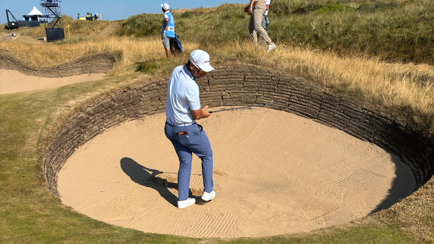 Lucas Herbert in a sand bunker, preparing to hit a shot. Two other people and grassy terrain are visible in the background under a clear sky.