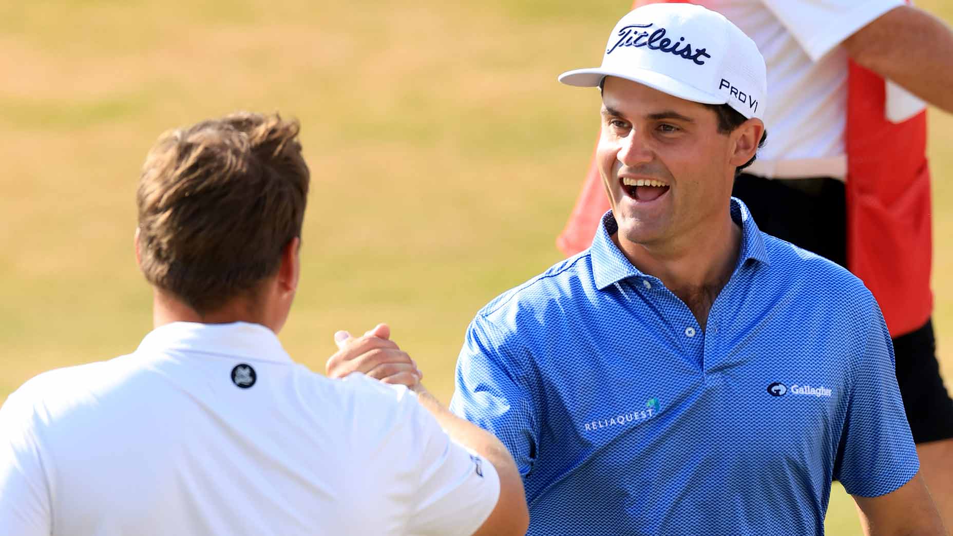 Two men on a golf course shake hands and smile. One wears a white shirt, while the other wears a blue shirt and a white Titleist cap. The background is out of focus.