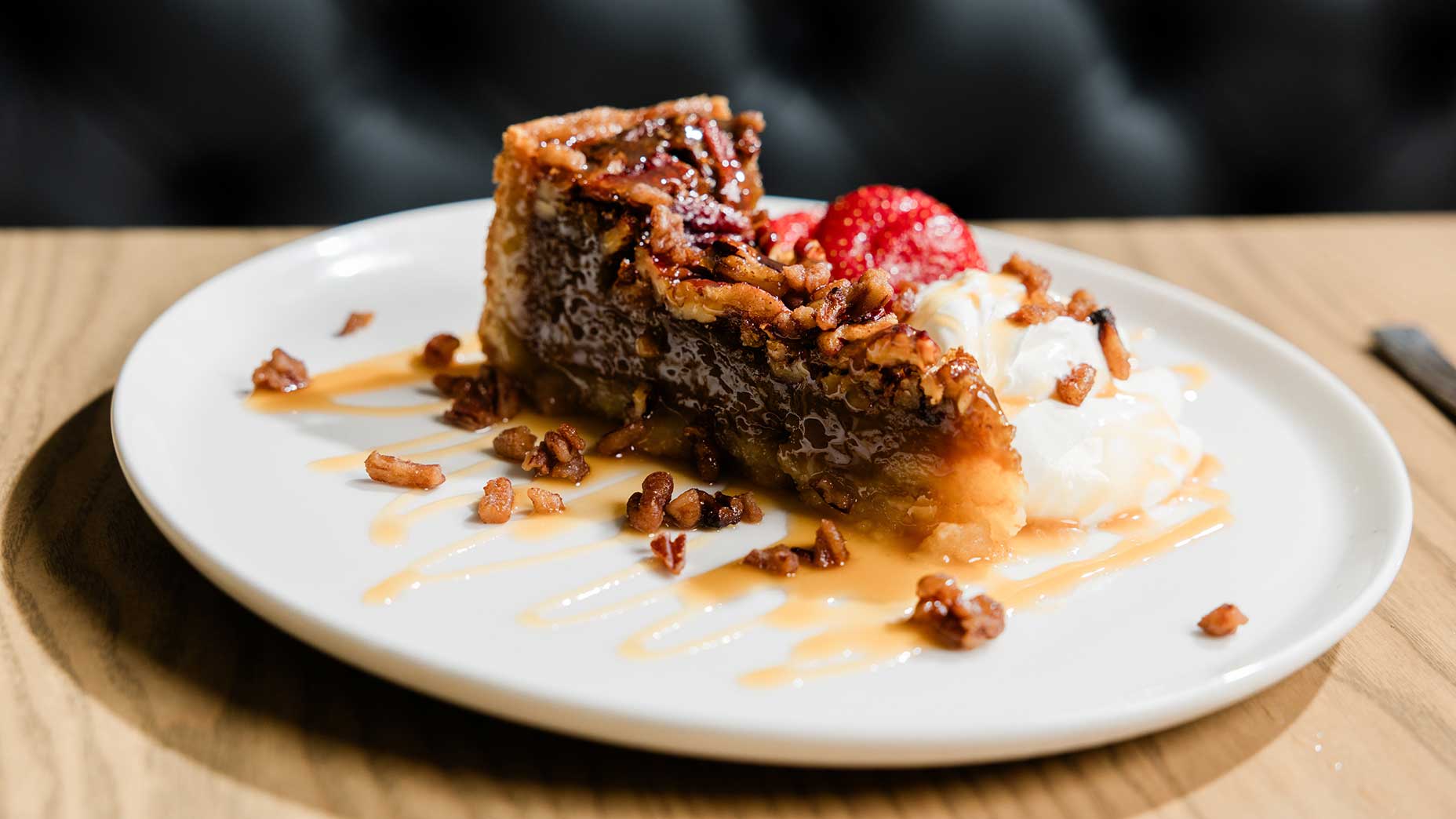A slice of pecan pie reminiscent of classic Southern and Scottish fare, topped with caramel sauce, chopped pecans, whipped cream, and strawberries, is artfully presented on a white plate set atop a wooden table—an indulgent treat worthy of McLemore Resort’s dessert menu.