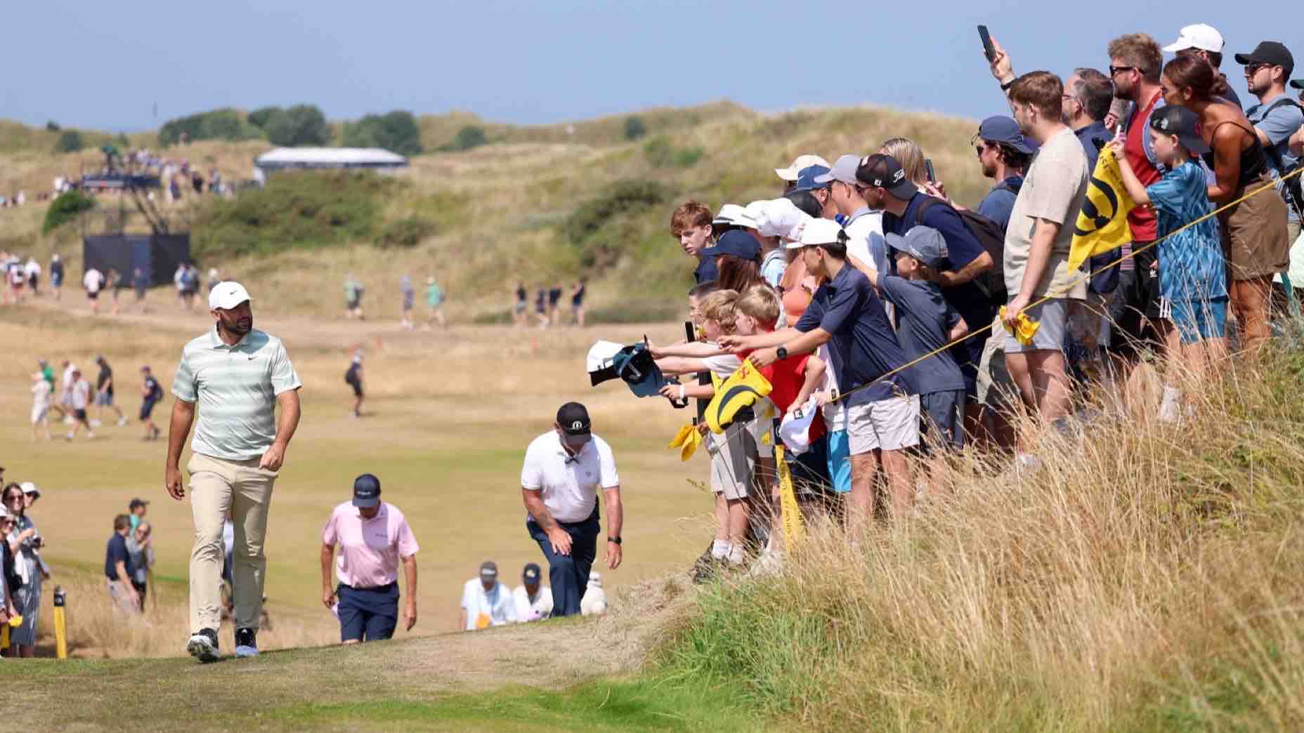 A golfer walks on a course while a large crowd of spectators stands nearby, some holding flags and phones. The scene is set on a grassy landscape under a clear sky.