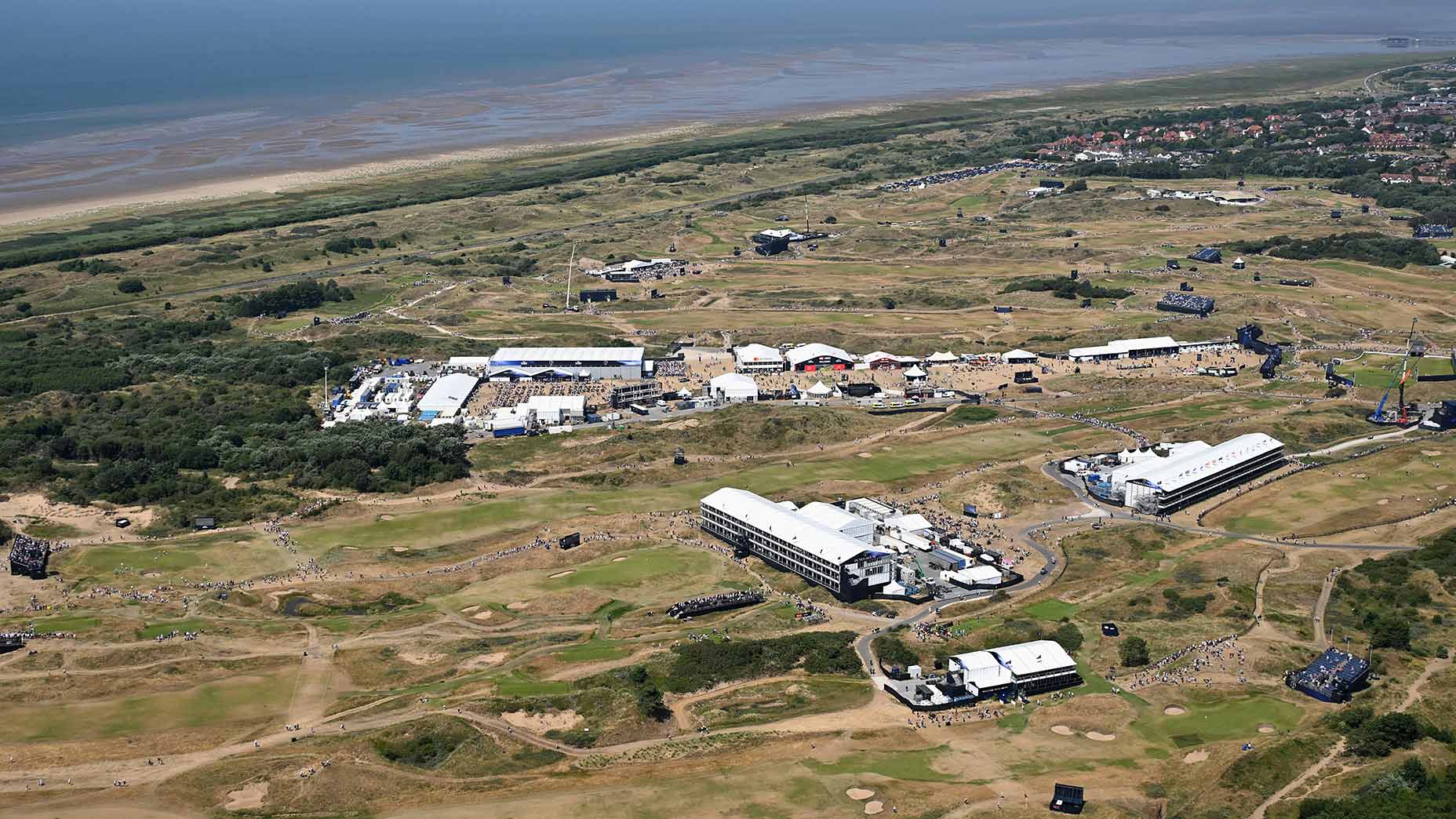 Aerial view of the renowned Royal Birkdale coastal golf course with several large white buildings, green fairways, sand traps, and the sea in the background. Scattered people and roads can be seen throughout the area, hinting at past Open Championship moments played on these storied grounds.