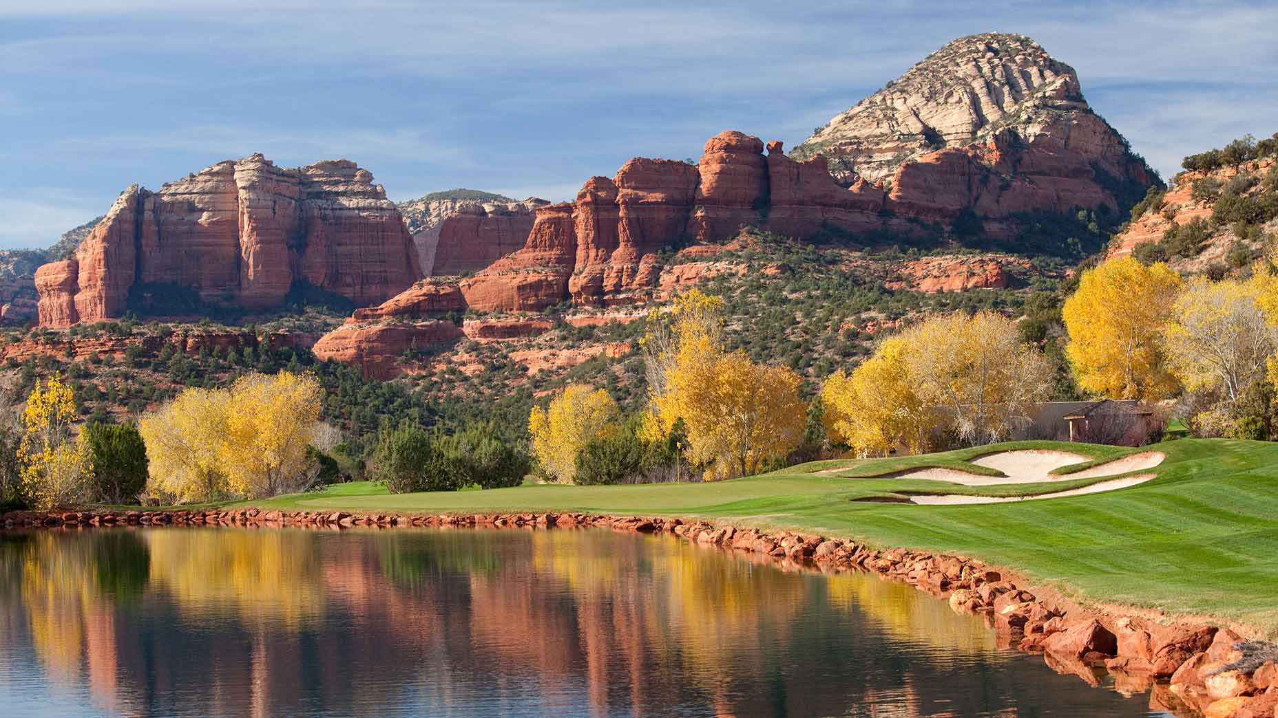 A golf course in Sedona, Ariz., with green grass and a sand trap sits by a calm lake, reflecting yellow-leaved trees and striking red rock mountains under a blue sky.