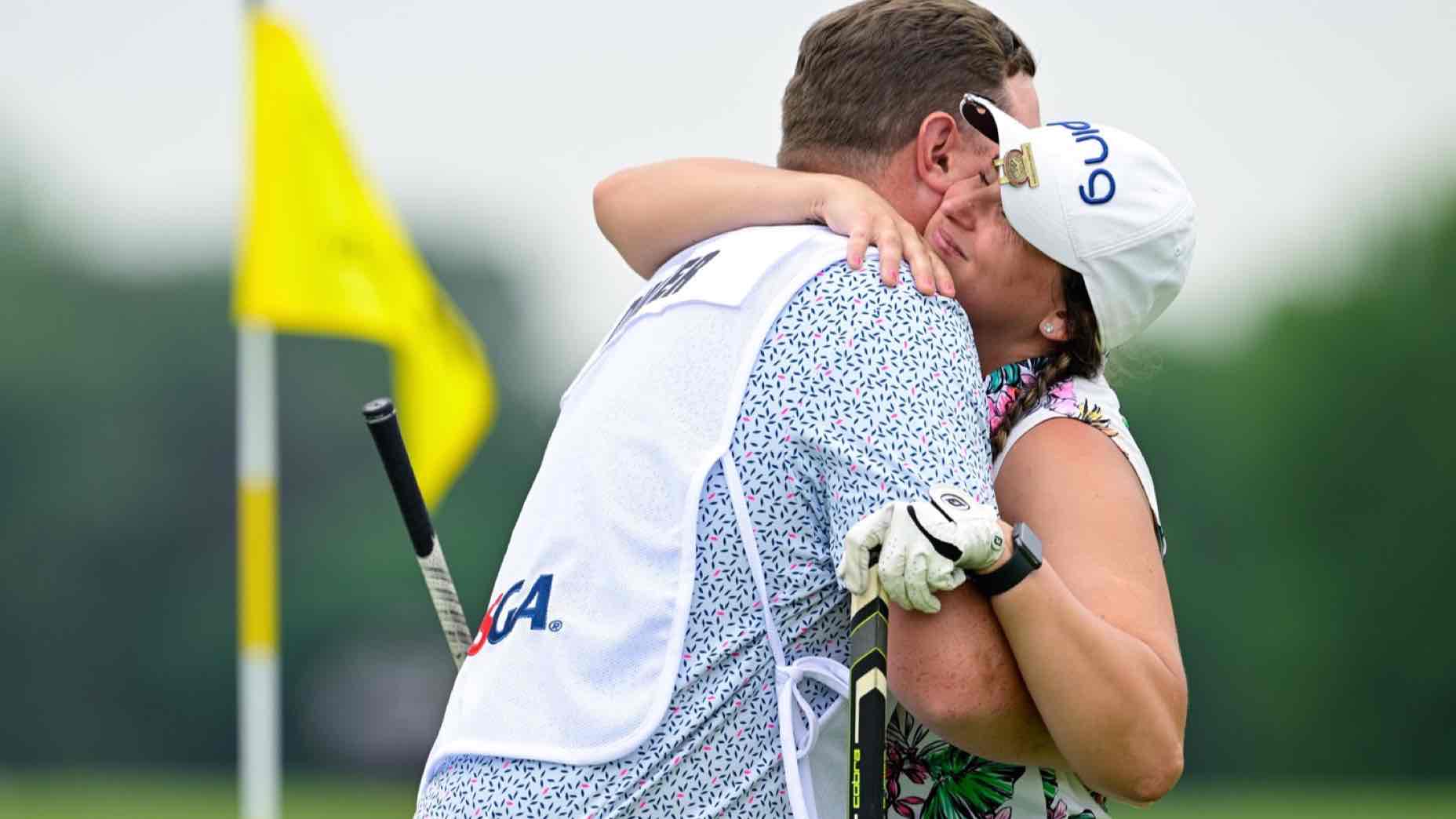 A golfer and her caddie share a joyful hug on a golf course, both smiling. This inspirational romance shines as the woman, holding a putter and wearing a cap and glove, stands by the yellow flag on the lush green grass in the background.