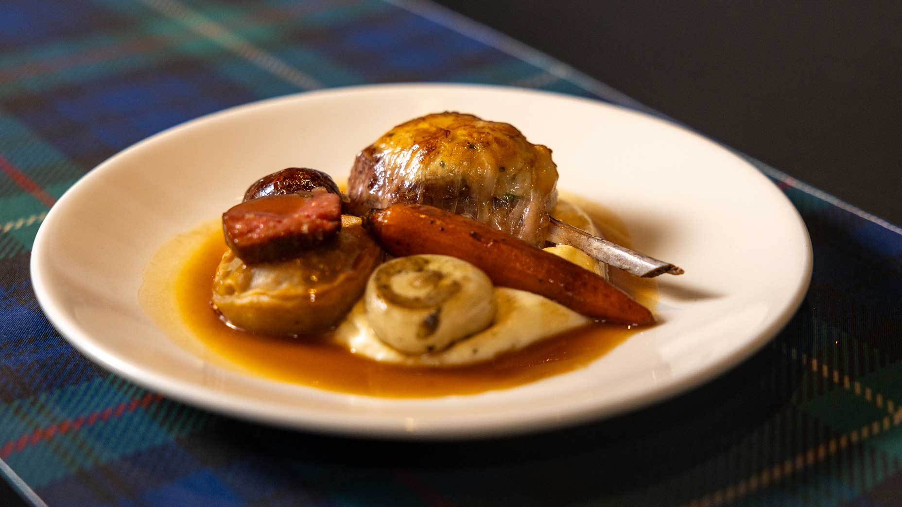 A white plate holds roasted meat, a carrot, mushrooms, a potato, and gravy, all served atop a bed of creamy mashed potatoes—an inviting showcase of Scottish fare—set on a blue tartan tablecloth.