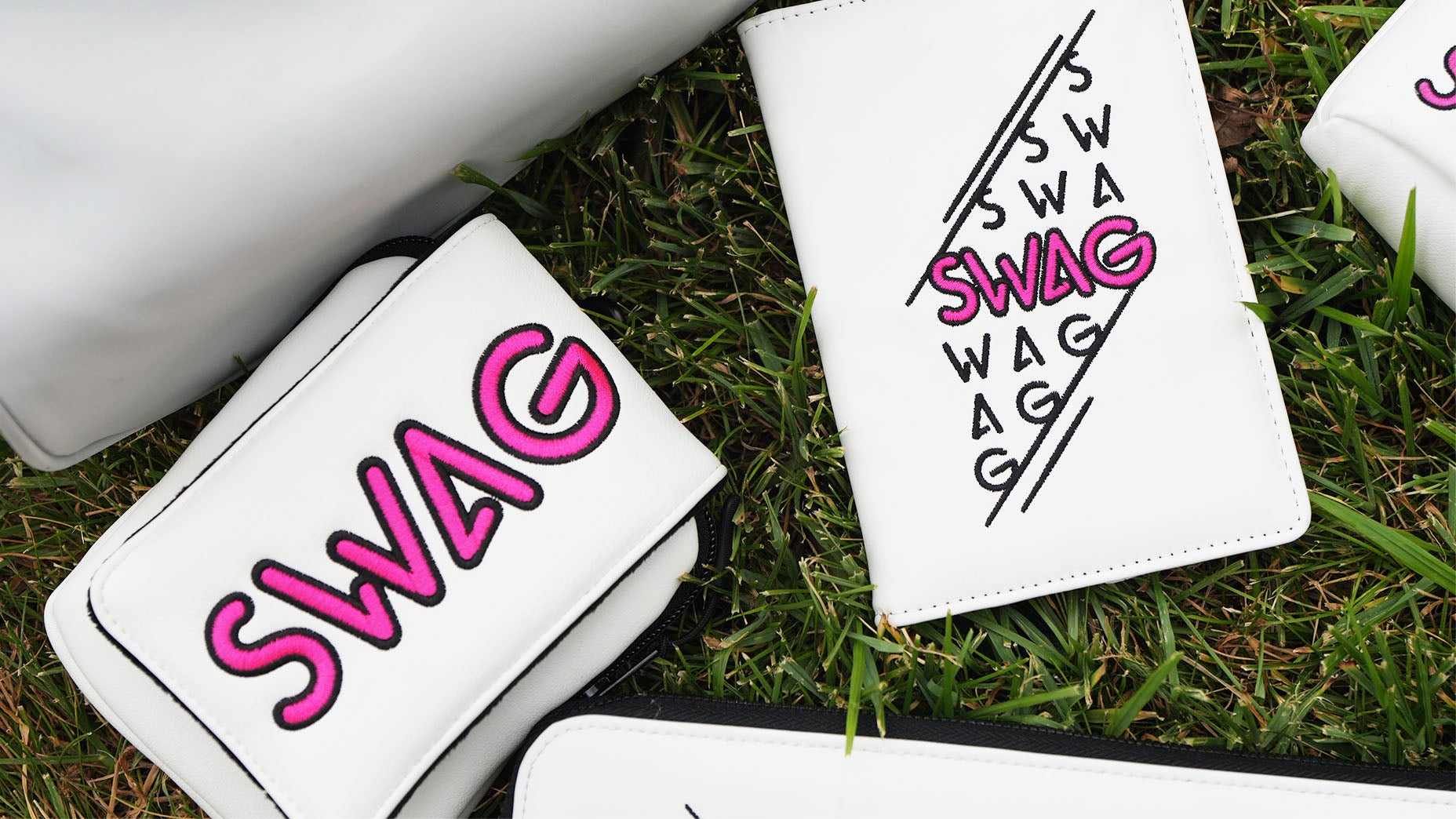 White wallets and a notebook with the word SWAG in bold pink and black letters are displayed on green grass, showcasing the latest SWAG accessories for your collection.