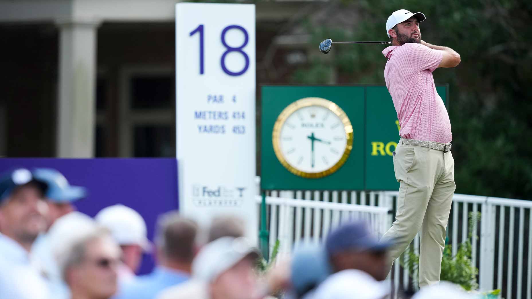 A golfer in a pink shirt and beige pants swings a club on the 18th hole during the 2026 FedEx St. Jude Championship Saturday tee times. A Rolex clock and a crowd of spectators are visible in the background. A sign shows the hole is a par 4 and 453 yards long.