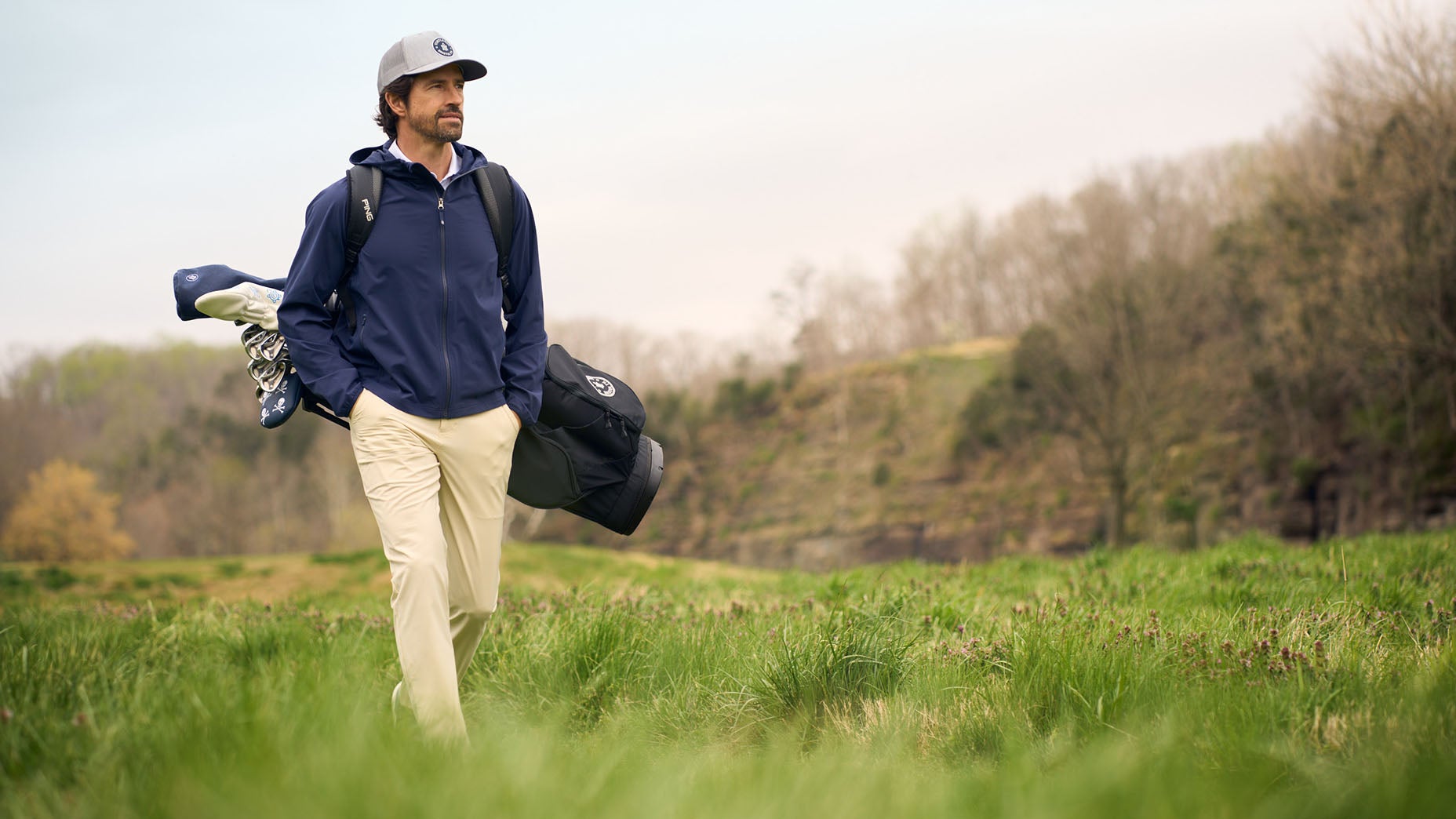 A man, layered in style with a cap and jacket from the Turtleson fall lineup, carries golf clubs over his shoulder while walking through a green grassy field with trees in the background.