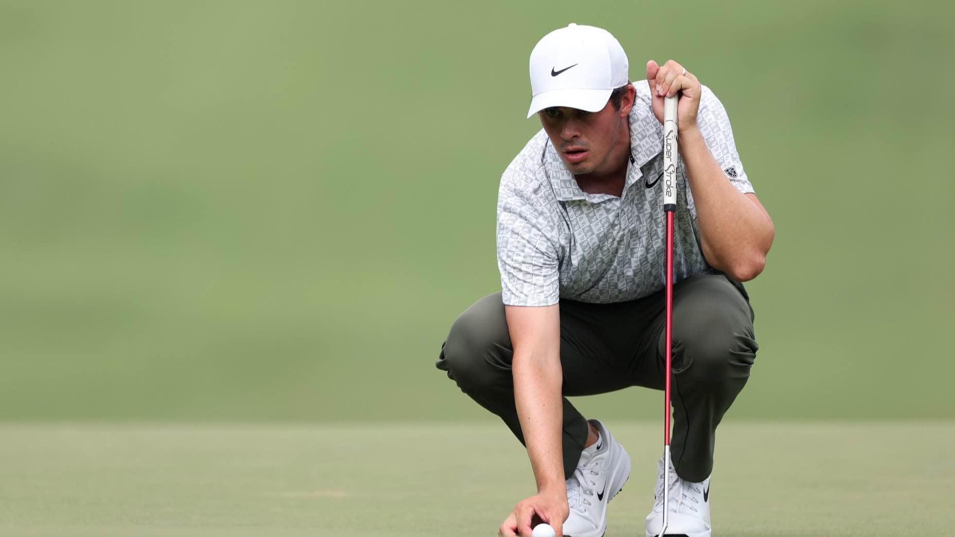 A golfer in a white cap and patterned shirt squats on the green, focusing on placing his golf ball while holding a putter, with a blurred grassy background.