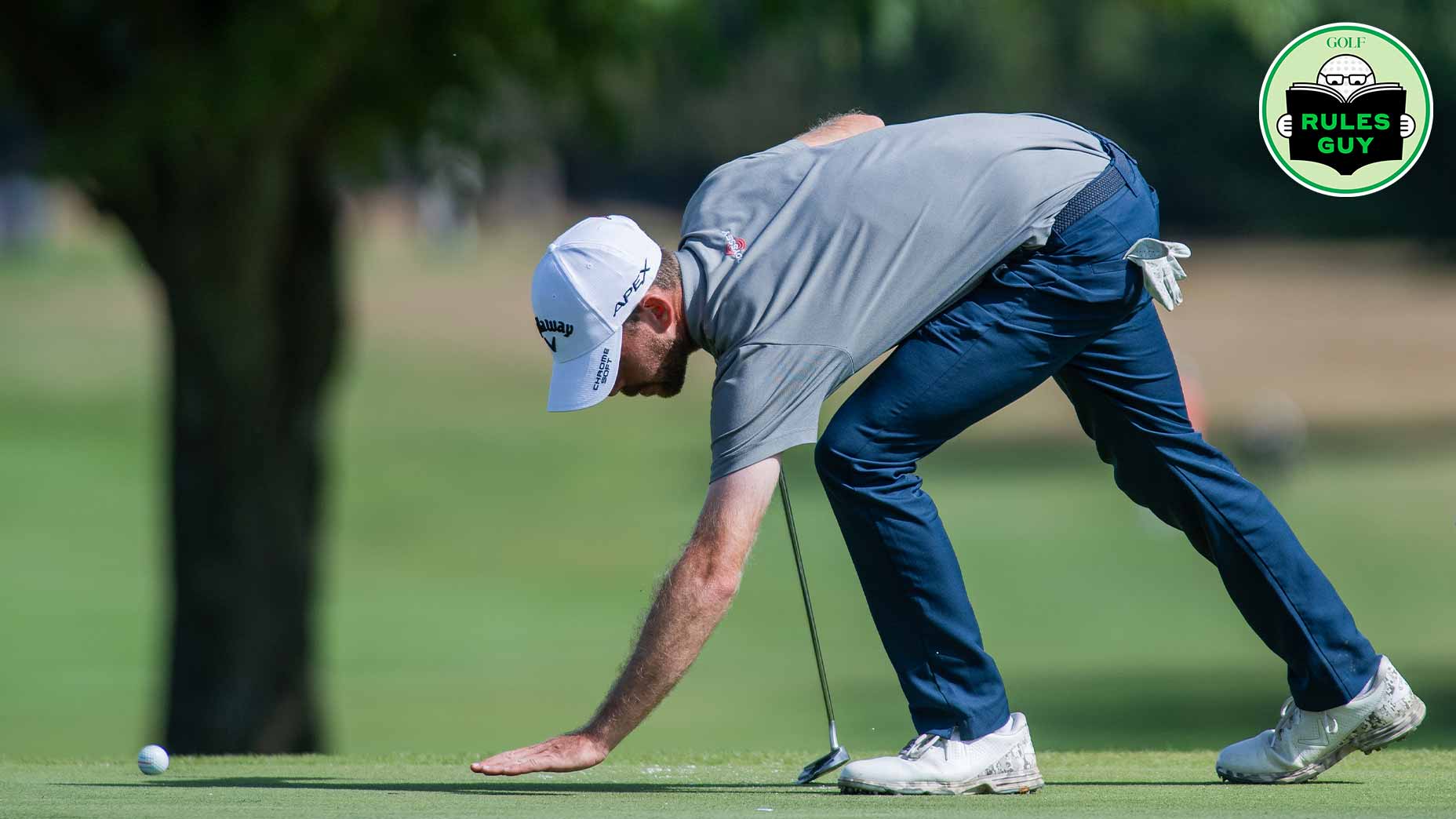 A golfer wearing a gray shirt, blue pants, and a white cap bends down to mark his golf ball on the green during a sunny day on the course, carefully mindful of the rules of golf as he checks for any loose impediments along his intended line. A Rules Guy badge appears in the top right corner of the image.