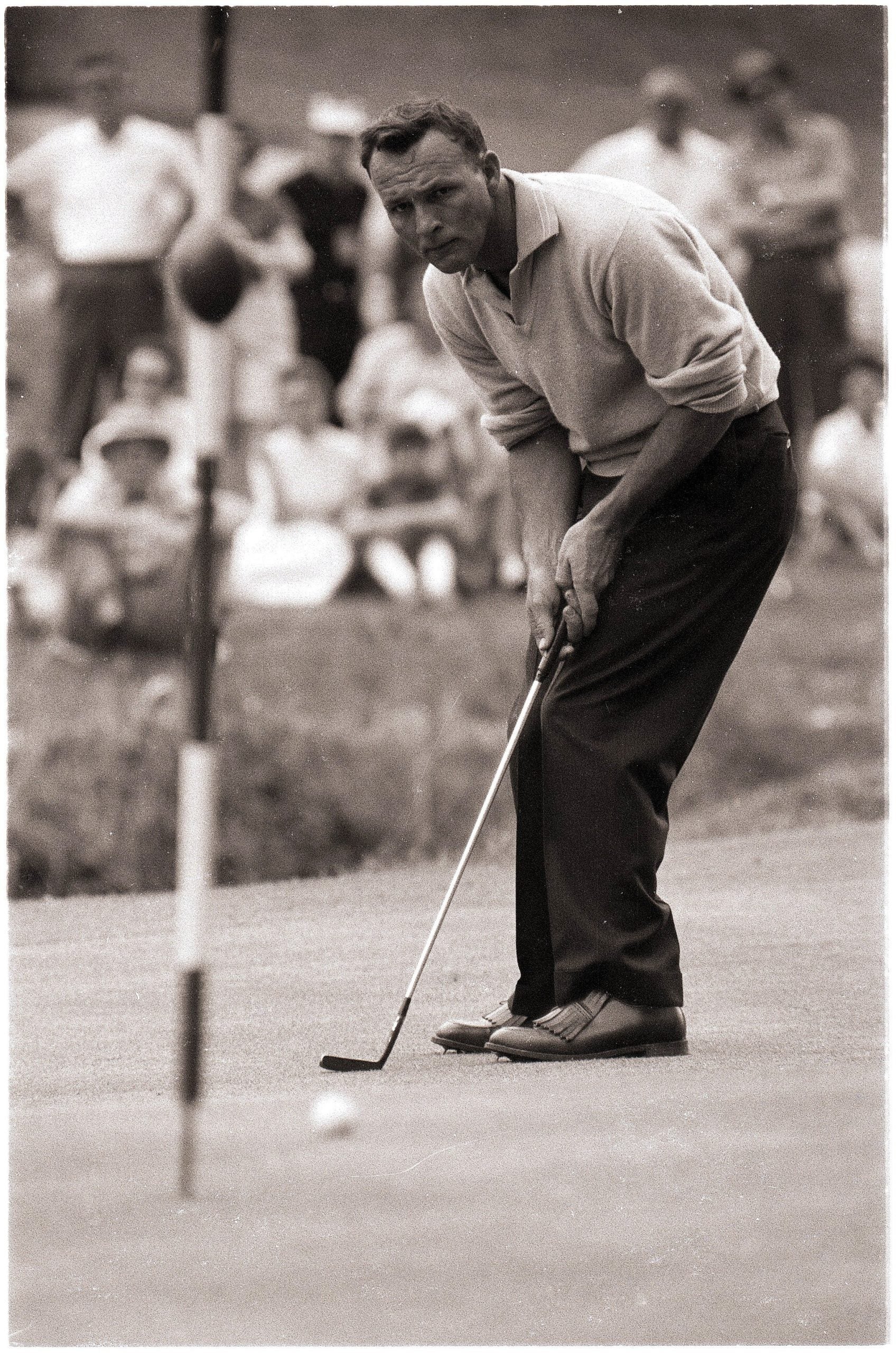 A man intently putts a golf ball on a green, with a crowd of people watching in the background. The image is black and white, and the golfer is wearing a sweater and dark pants.